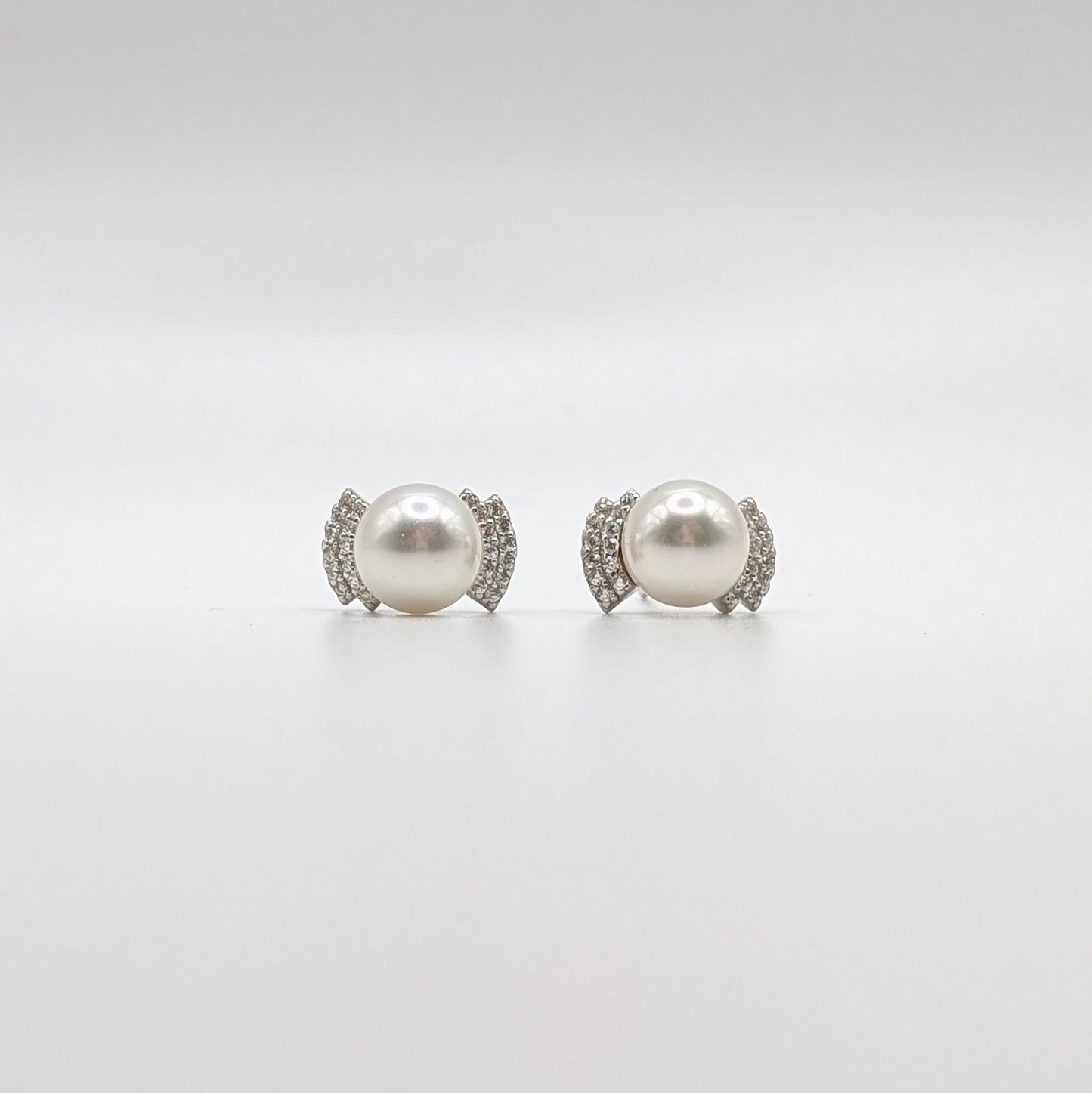 A pair of stud earrings featuring a large white pearl in a silver-toned setting accented with small clear stones, photographed on a seamless light gray background.
