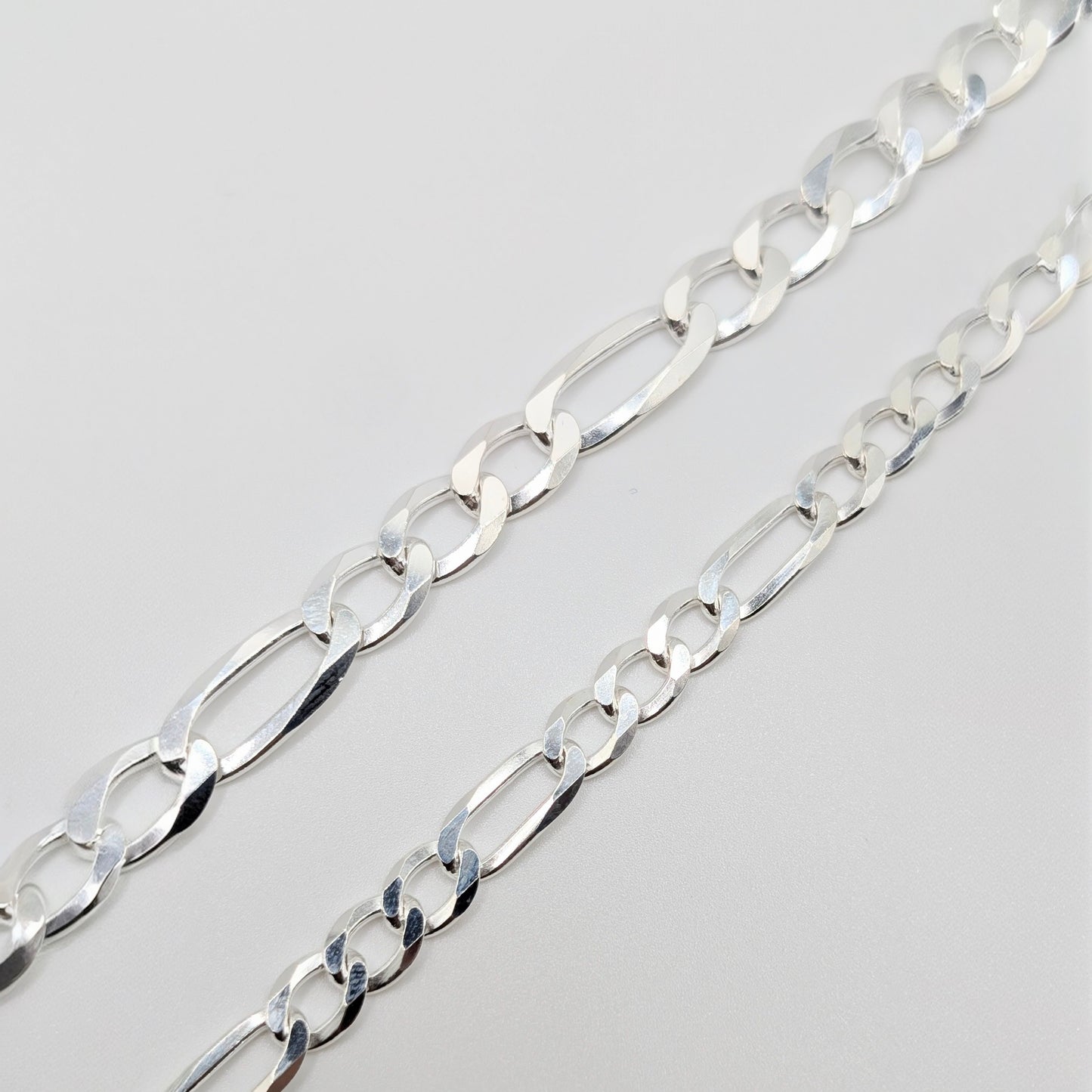 A close-up of a polished silver Figaro chain bracelet, showing the interlocking oval links.