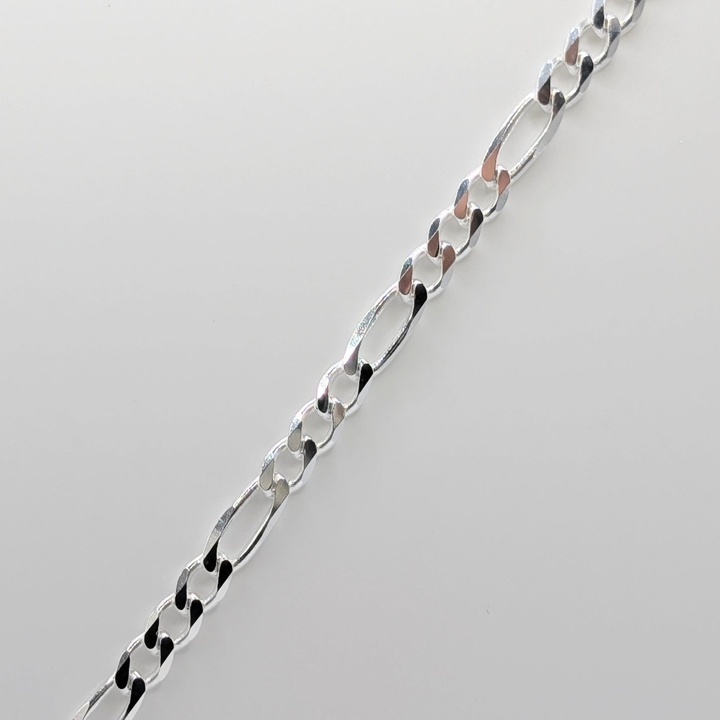 A close-up of a polished silver Figaro chain necklace laid diagonally on a light gray background.