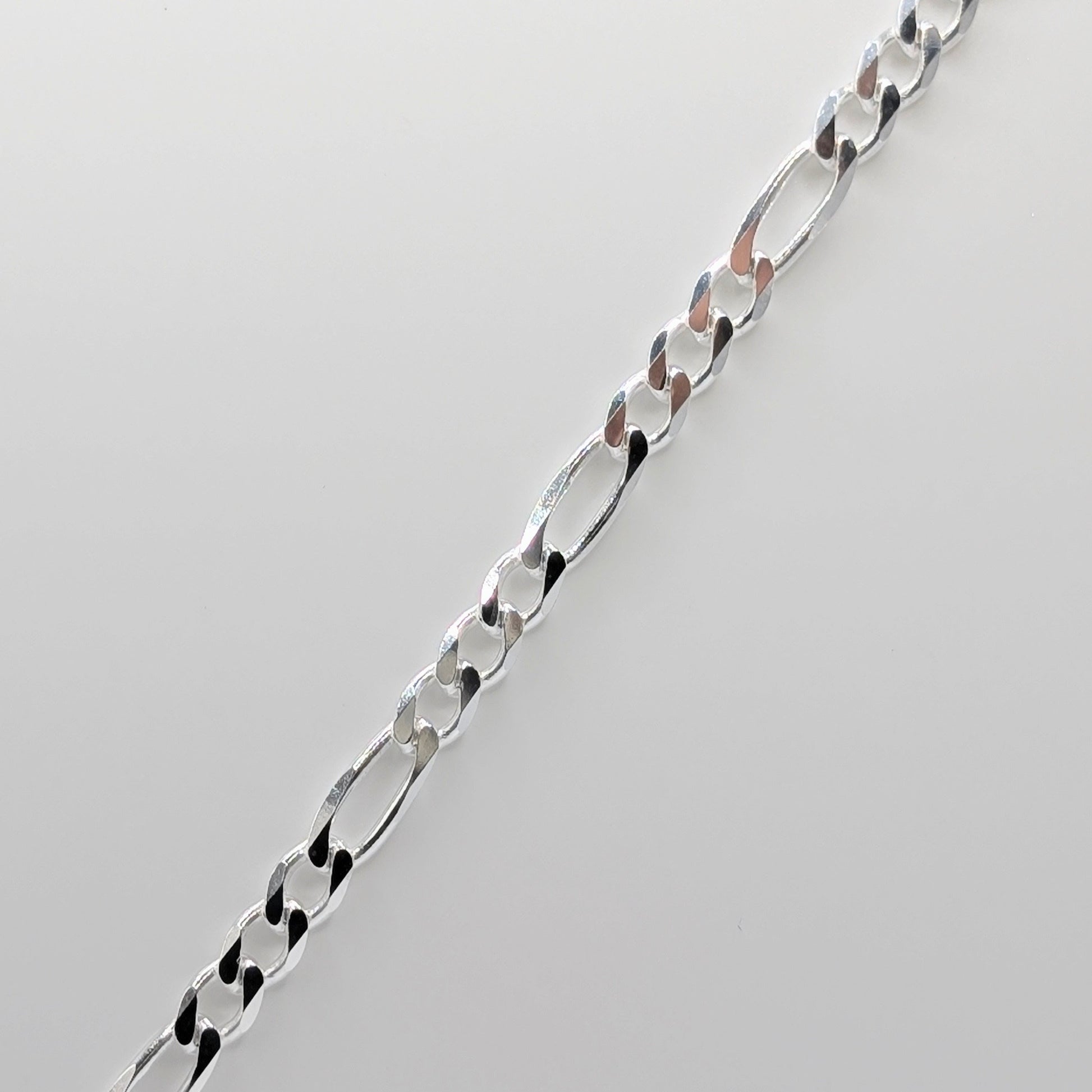 A close-up of a polished silver Figaro chain necklace laid diagonally on a light gray background.