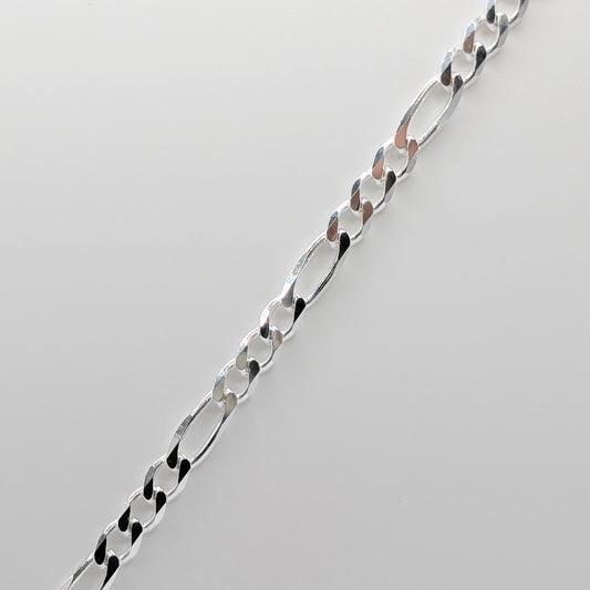 A close-up of a polished silver Figaro chain necklace laid diagonally on a light gray background.