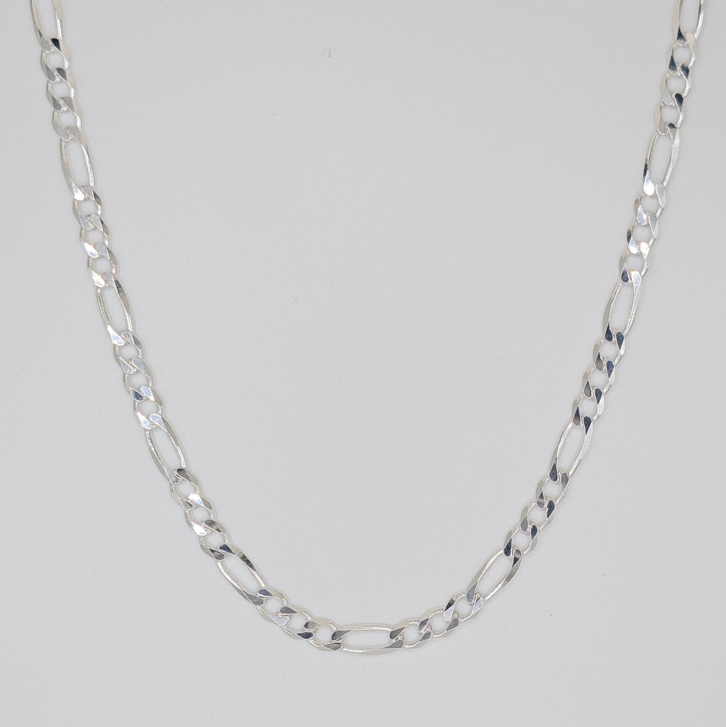 A silver Figaro chain necklace with alternating oval and elongated links, displayed in a U-shape against a neutral light gray background.