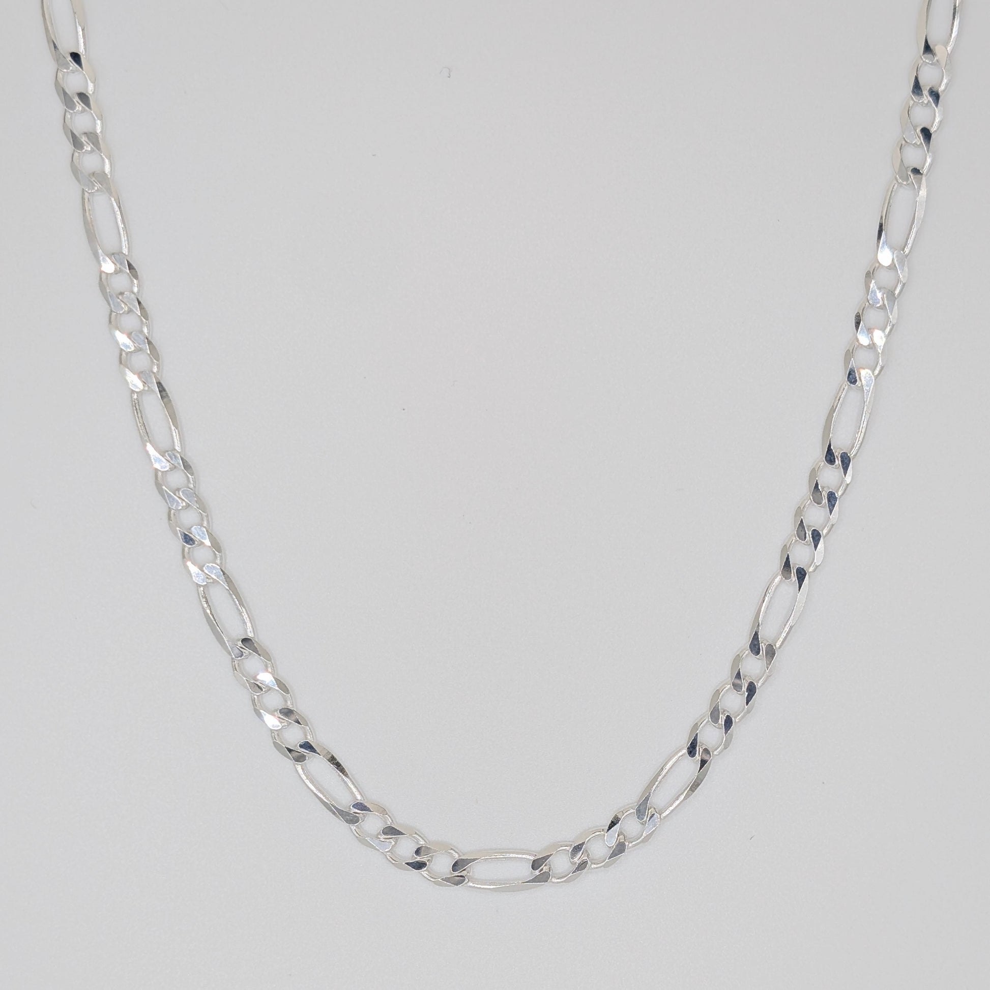 A silver Figaro chain necklace with alternating oval and elongated links, displayed in a U-shape against a neutral light gray background.