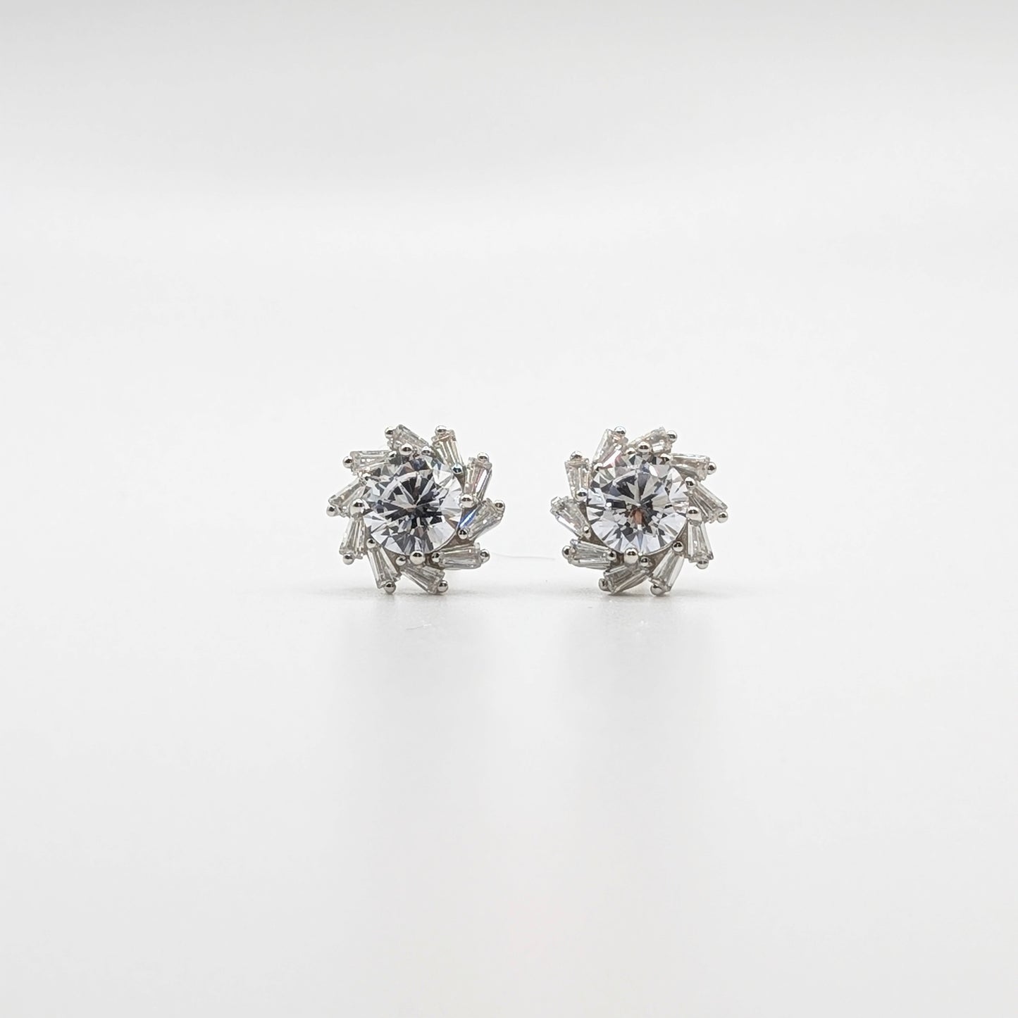 A pair of silver-toned stud earrings with a starburst cluster design featuring clear baguette and round-cut stones, set against a seamless white background.