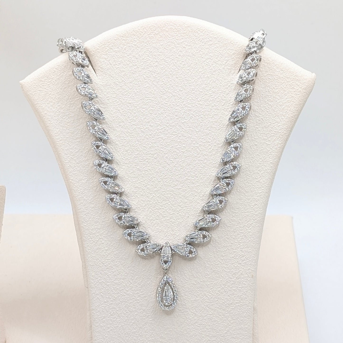 A sparkling silver-tone diamond necklace displayed on a cream-colored jewelry bust. The necklace features marquise-cut and round-cut diamonds set in a silver-toned metal, with a teardrop-shaped diamond pendant at the center.
