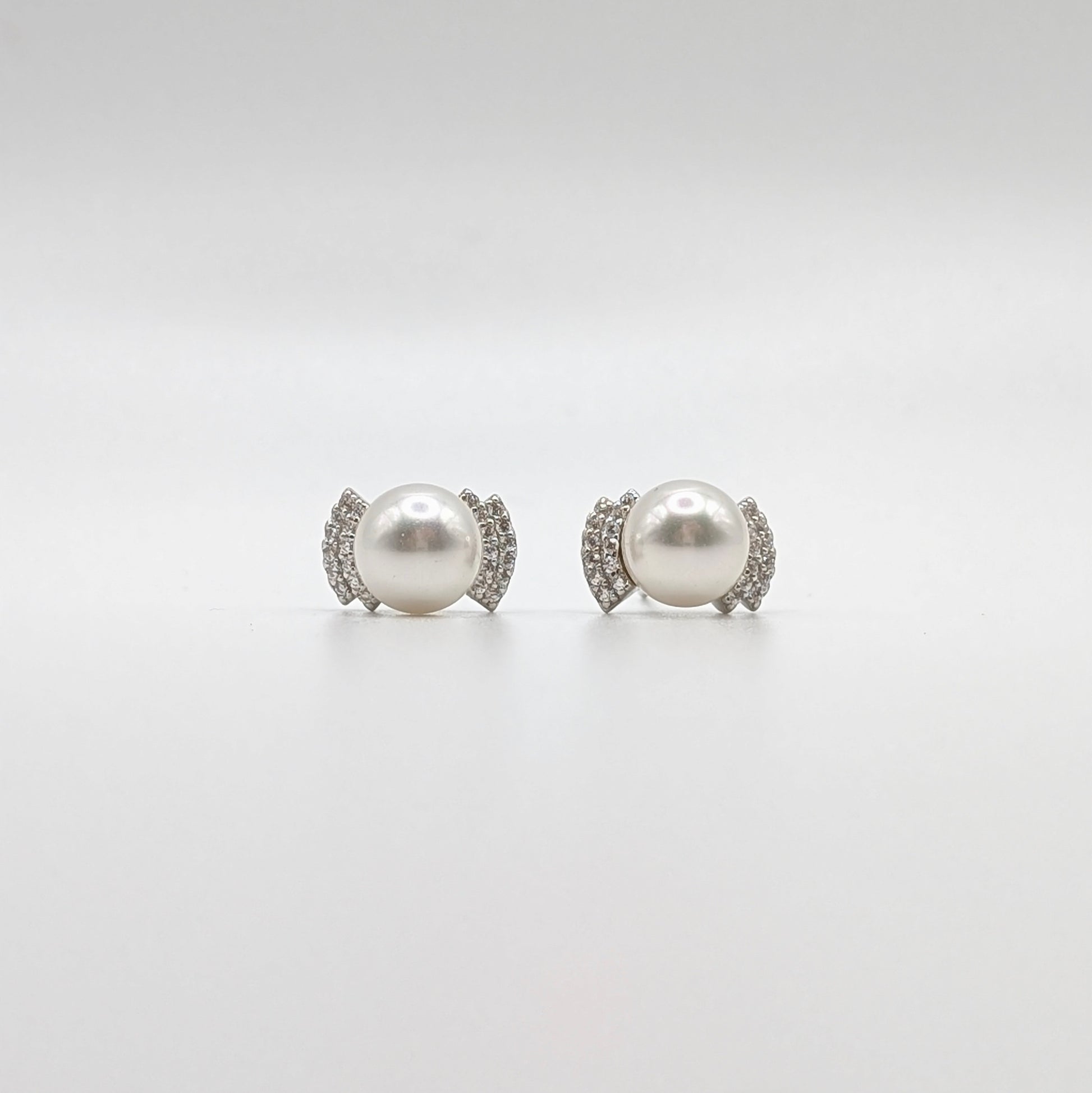 A pair of stud earrings featuring a large white pearl in a silver-toned setting accented with small clear stones, photographed on a seamless light gray background.
